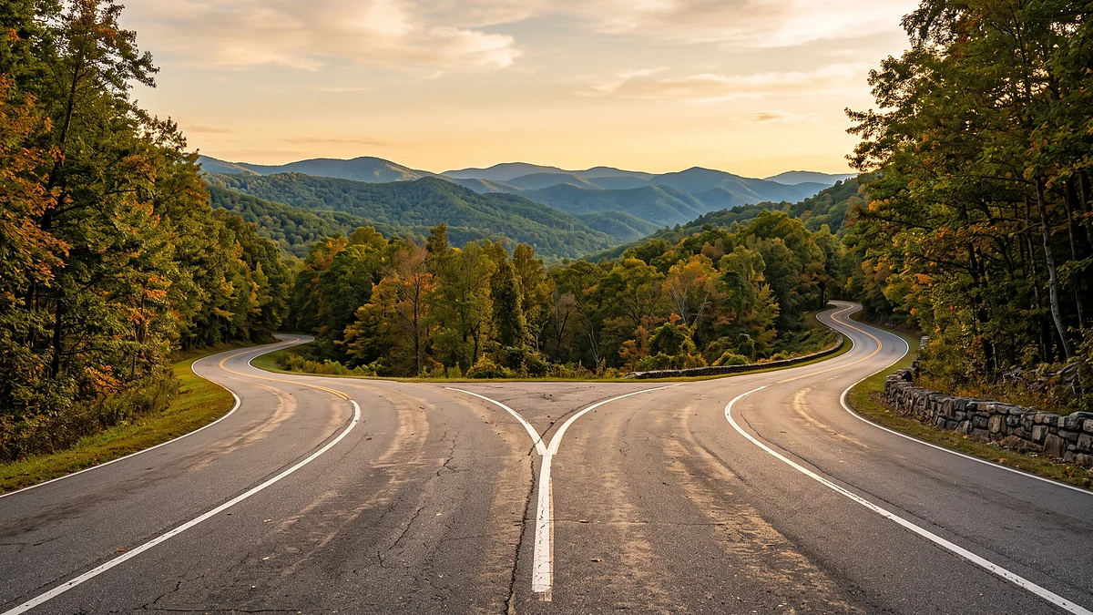A two-lane mountain road splitting into a fork, one branch curving left and the other curving right, seen low from the centre line at golden hour. Layered forested ridgelines recede toward a pale gold sky, with early-autumn hardwoods on both shoulders and a low dry-stone wall on the right.