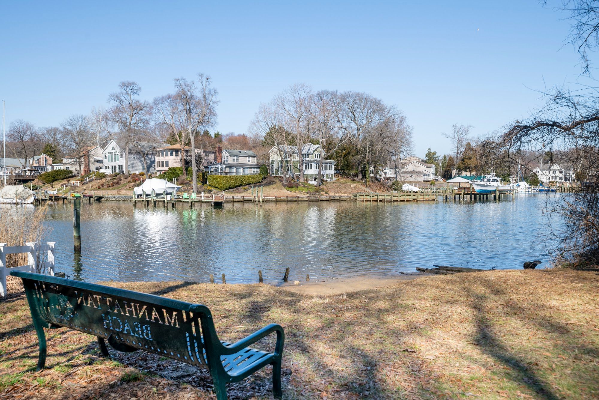 A green Manhattan Beach bench on a community beach, with homes and boats across the creek