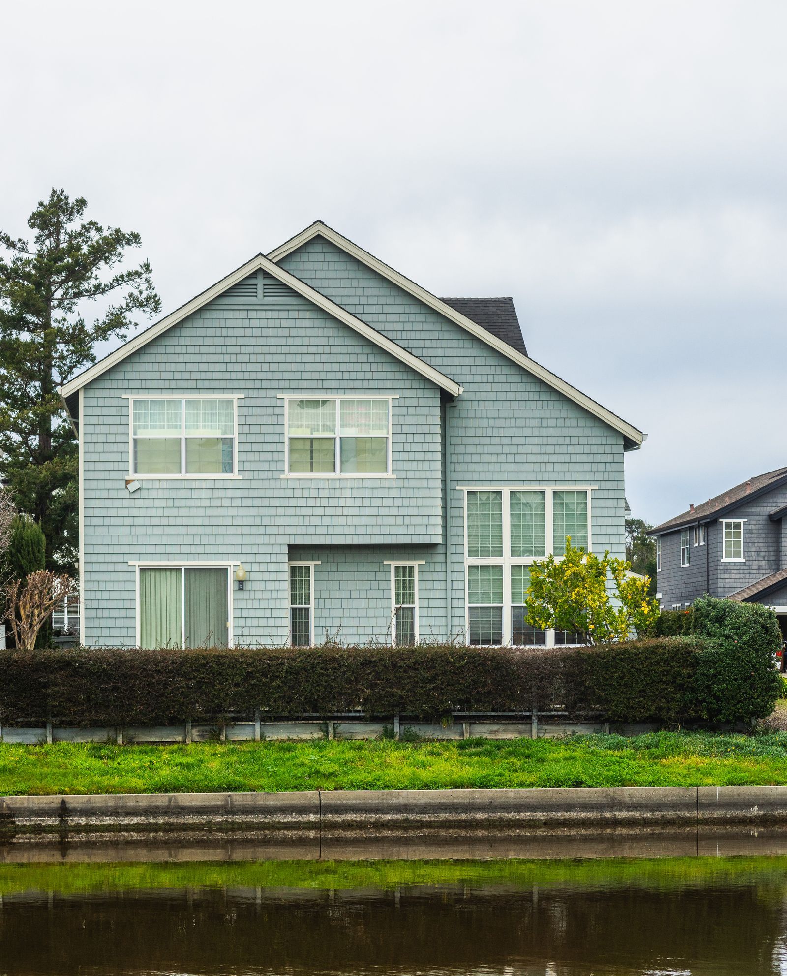 Grey shake-sided detached family home behind a hedge beside water, typical of Bronte or southwest Oakville