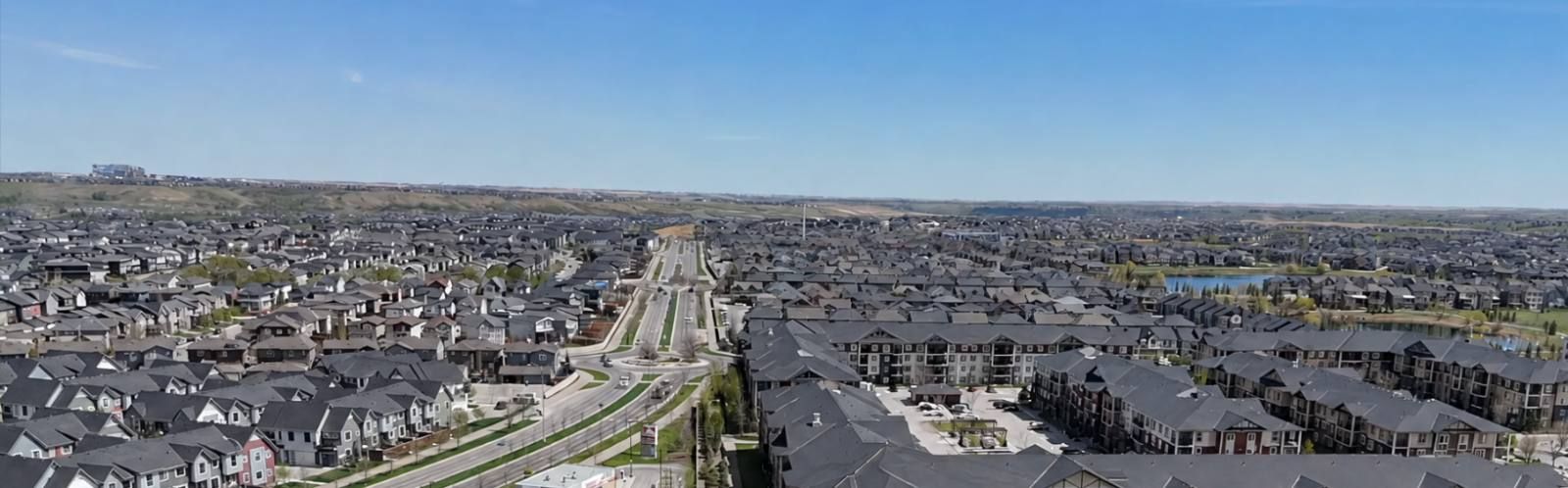 Aerial view of Legacy, Calgary, with a tree-lined boulevard, townhouses and low-rise apartment buildings stretching toward a stormwater pond and the prairie horizon