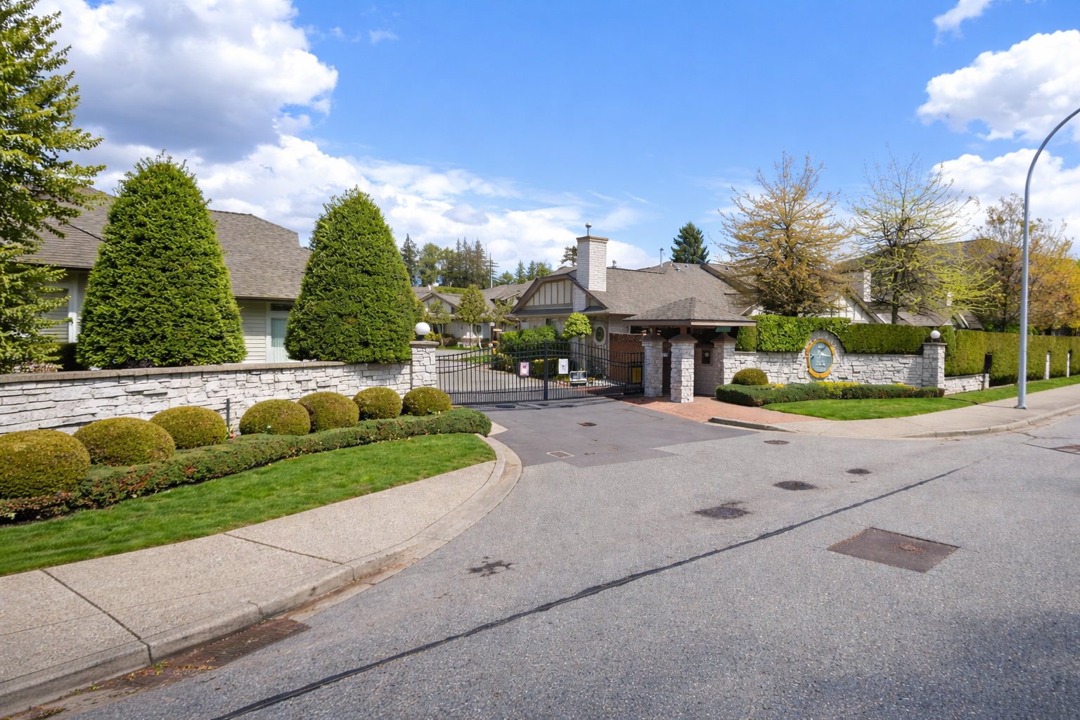 Street-level view of Hampton Woods gated townhouse entrance at 16325 82 Avenue in Fleetwood Surrey, showing landscaped entry, stone wall, and residential buildings on a clear day