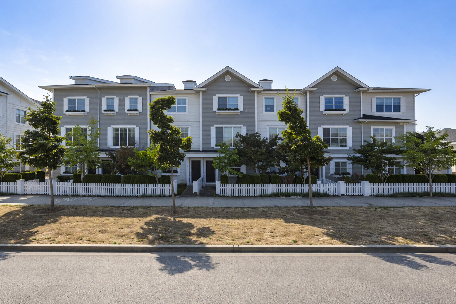 Street-facing view of Pier 16 townhomes in South Surrey showing modern grey and white multi-level units with white picket fencing, landscaped trees, and clear blue sky.