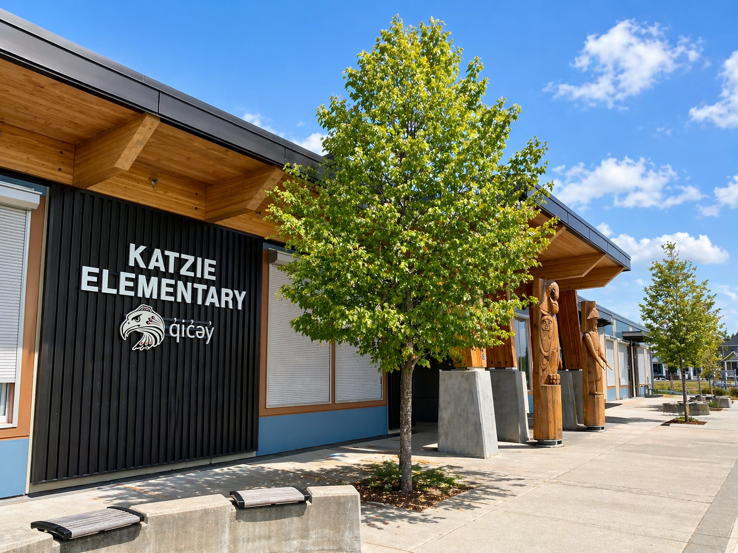 Exterior of Katzie Elementary School in Clayton Heights Surrey BC featuring modern building design, wood accents, carved totem poles, and landscaped walkway under a clear sky.