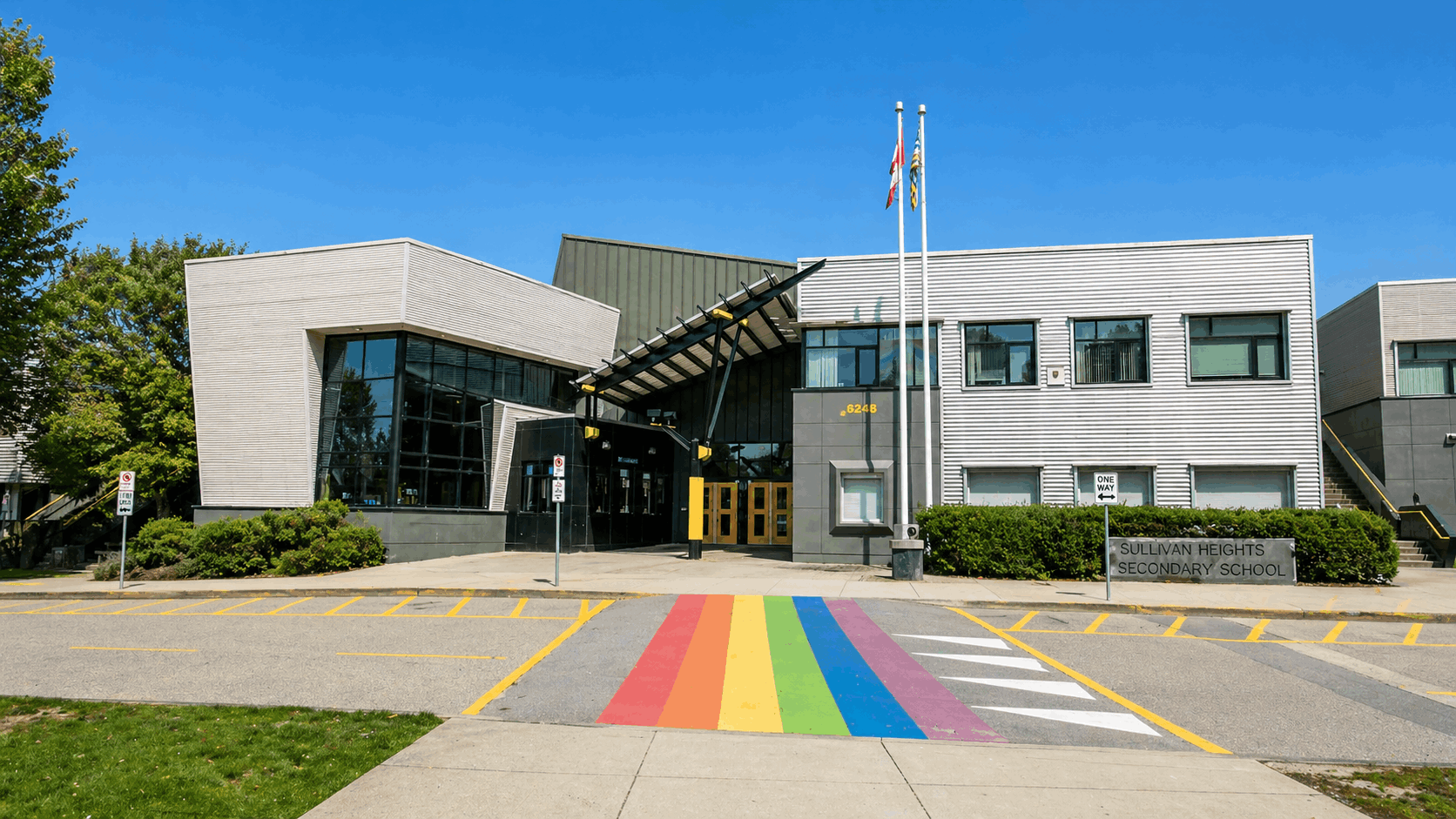 Front exterior view of Sullivan Heights Secondary School in Surrey BC, featuring modern architecture, glass entryway, flags, and a rainbow crosswalk in the Sullivan Heights neighbourhood of Newton.