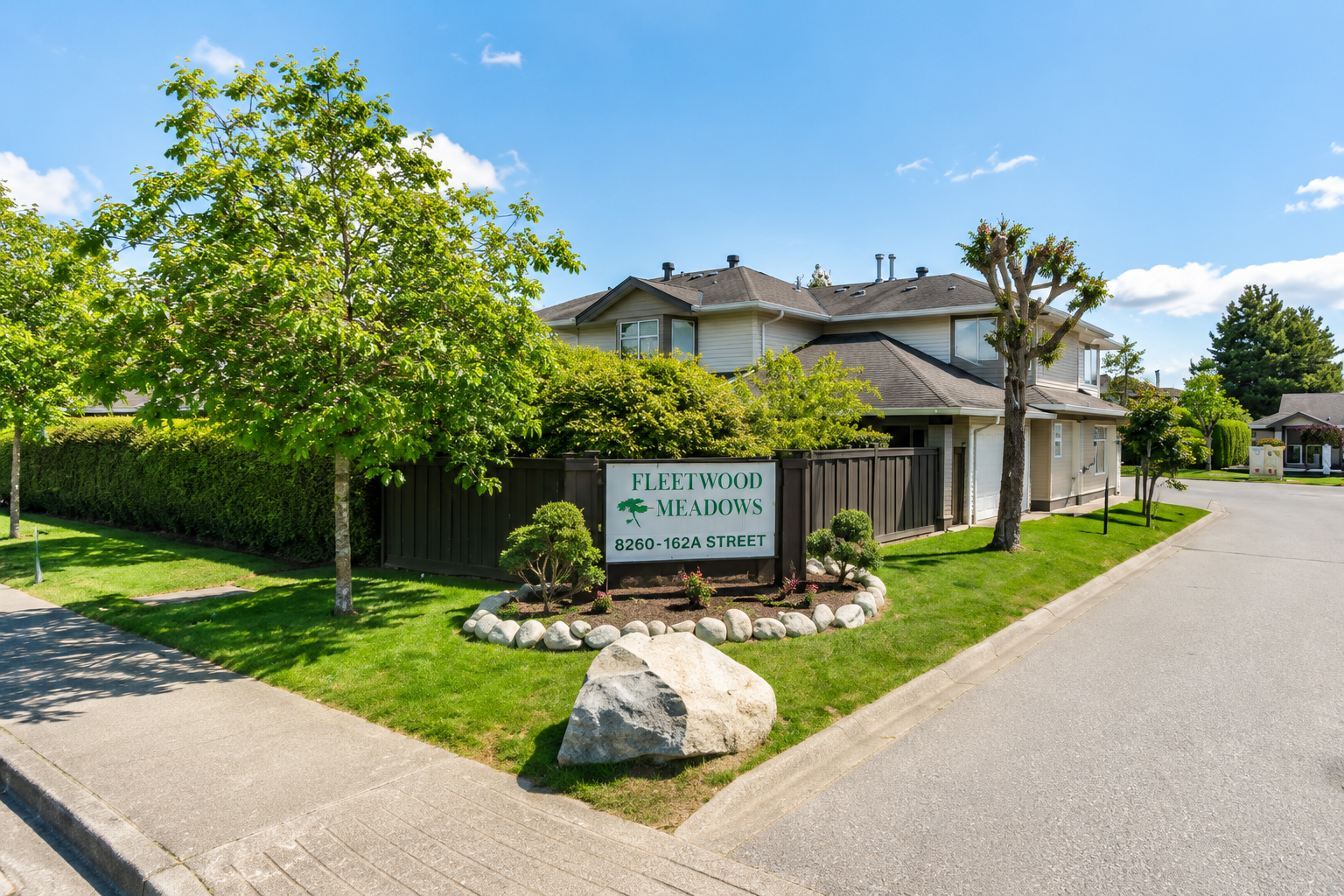 Main entrance sign for Fleetwood Meadows Townhomes at 8260 162A Street in Surrey BC featuring landscaped grounds, mature trees, green lawns, townhouse buildings, and the entrance roadway into the Fleetwood community.