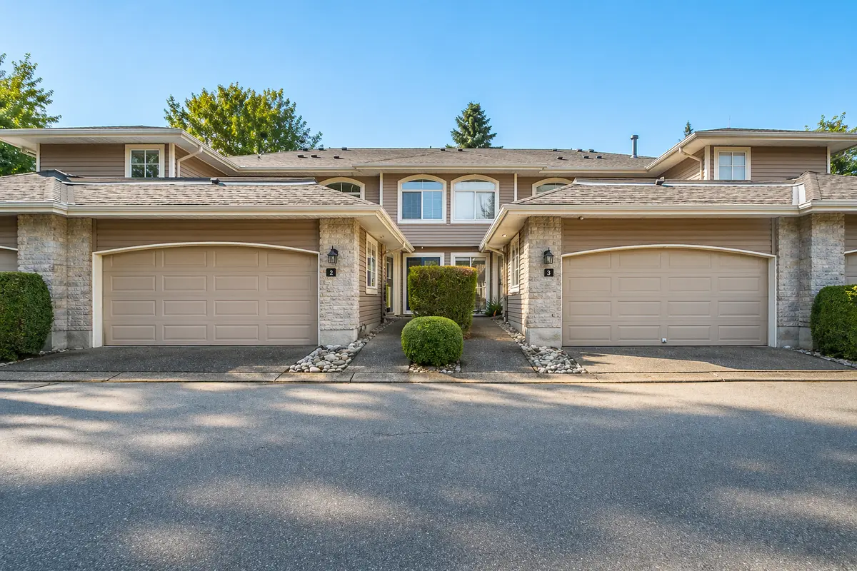 Exterior view of Peninsula Village townhomes in South Surrey, BC, showing double garage fronts, stone accents, landscaped entry walkway, and two-level townhouse design in a 55+ gated community.