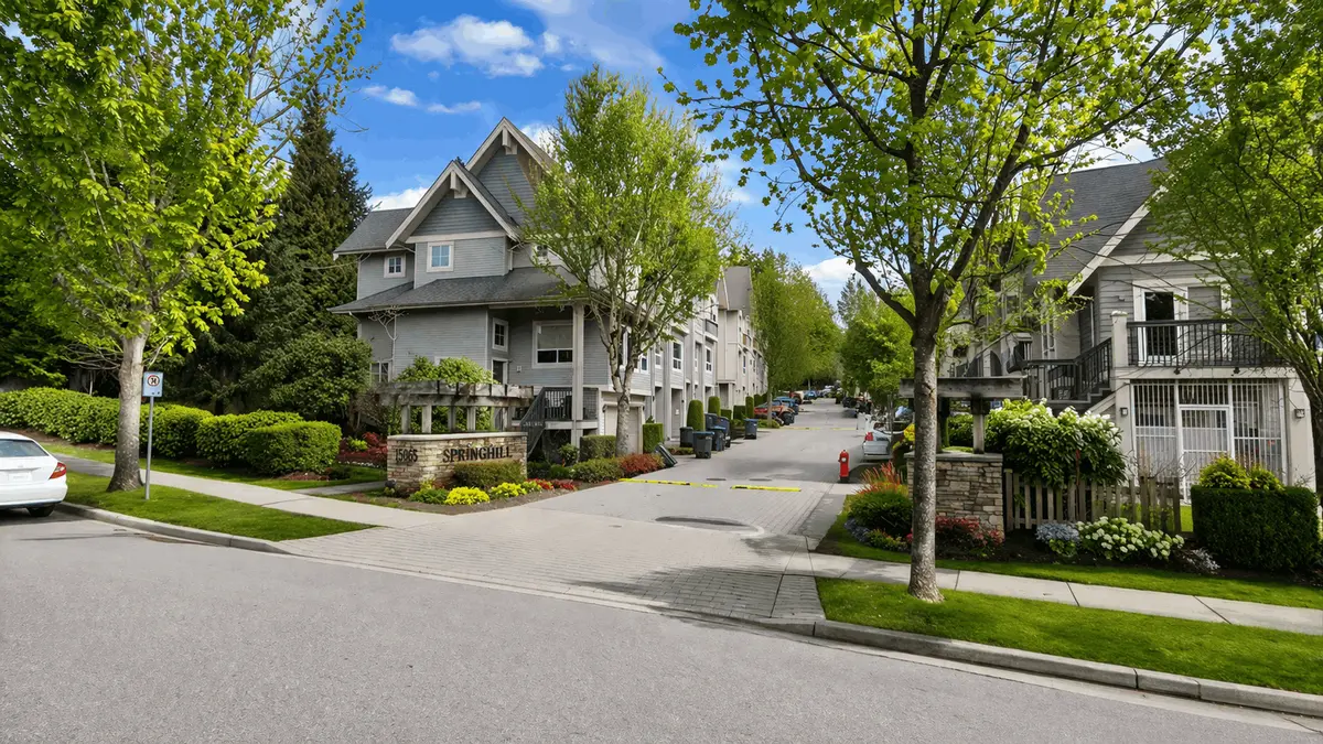 Street-level view of Springhill townhomes at 15065 58 Avenue in Surrey BC showing entrance signage, blue sky, and vibrant green landscaping from the road
