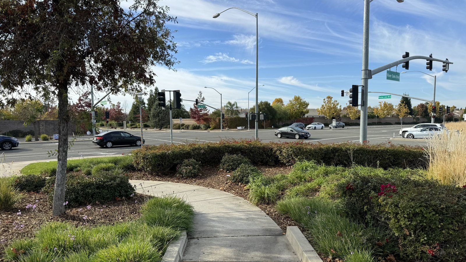Wide landscaped intersection of Plumas Lake Boulevard and River Oaks Boulevard in Plumas Lake, California, with a walking path, shrubs, trees, and traffic signals under a blue sky