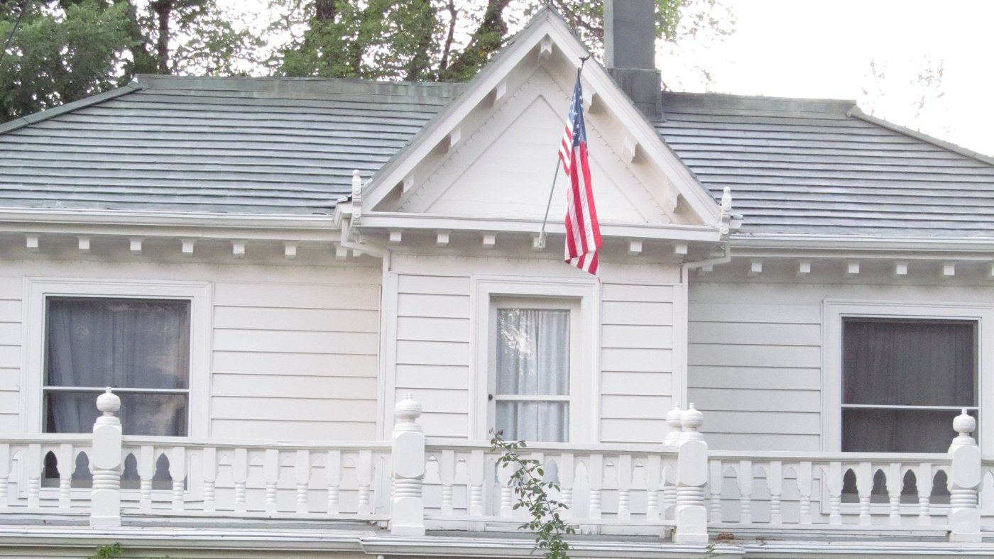 White two-story historic home with a gabled roof, wide second-floor balcony with turned balusters, and tall trees behind it in Nevada City, California