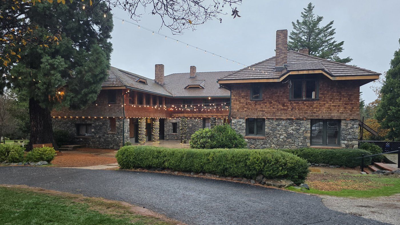Historic stone and wood-shingle North Star House in Grass Valley, California, with tall pines and a large sequoia, trimmed hedges, and a curving paved driveway