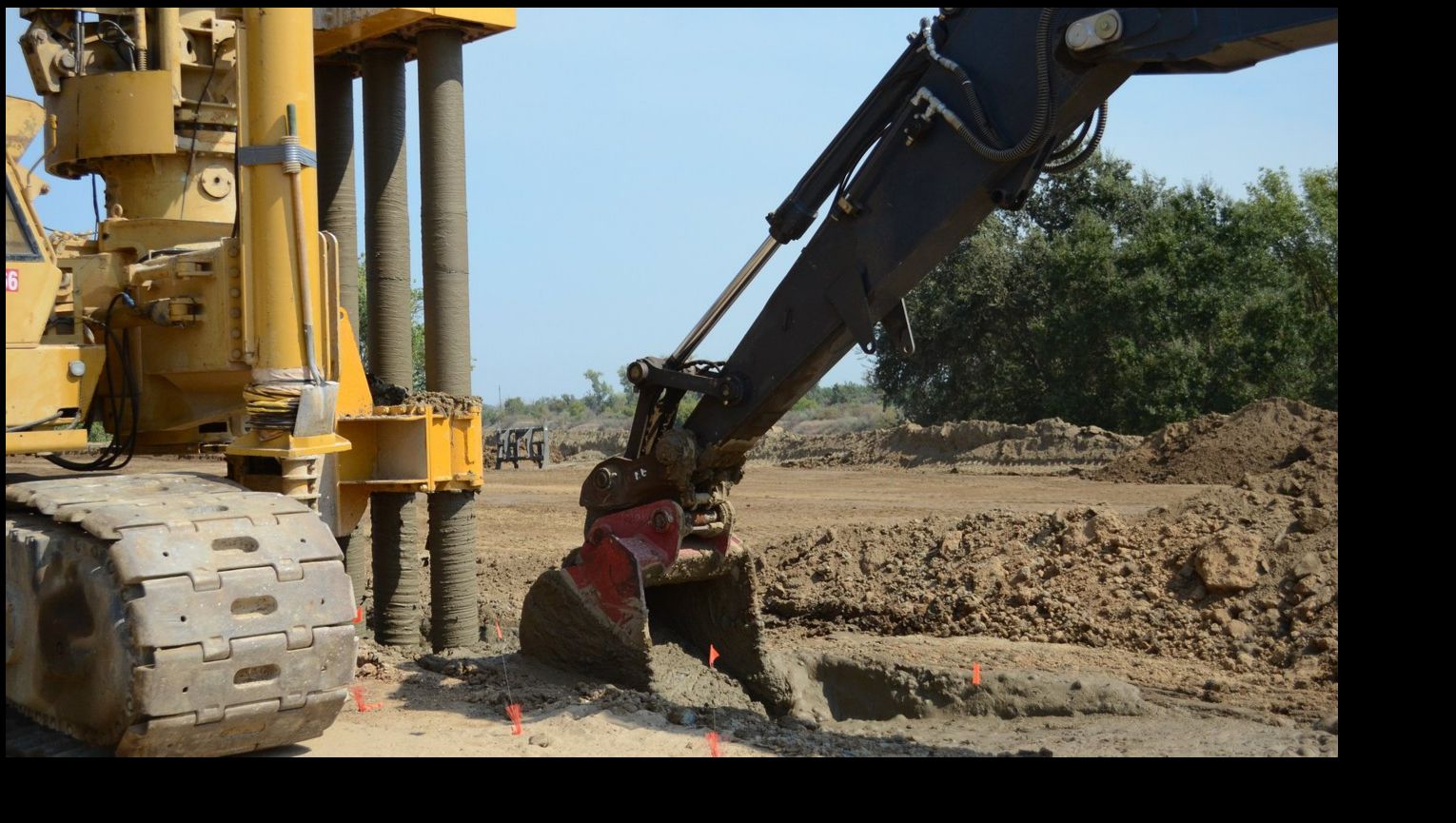Heavy drilling rig and excavator building an underground slurry wall in a cleared dirt work zone along the Feather River levee at Shanghai Bend in Yuba City, California, with green trees in the background