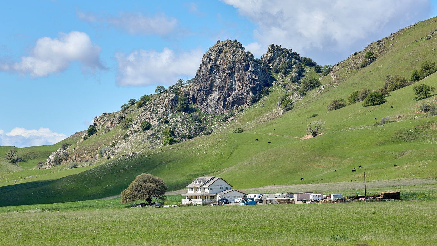 White two-story farmhouse with outbuildings on a green pasture at the base of the rocky Sutter Buttes under a blue sky with scattered clouds in Sutter County, California