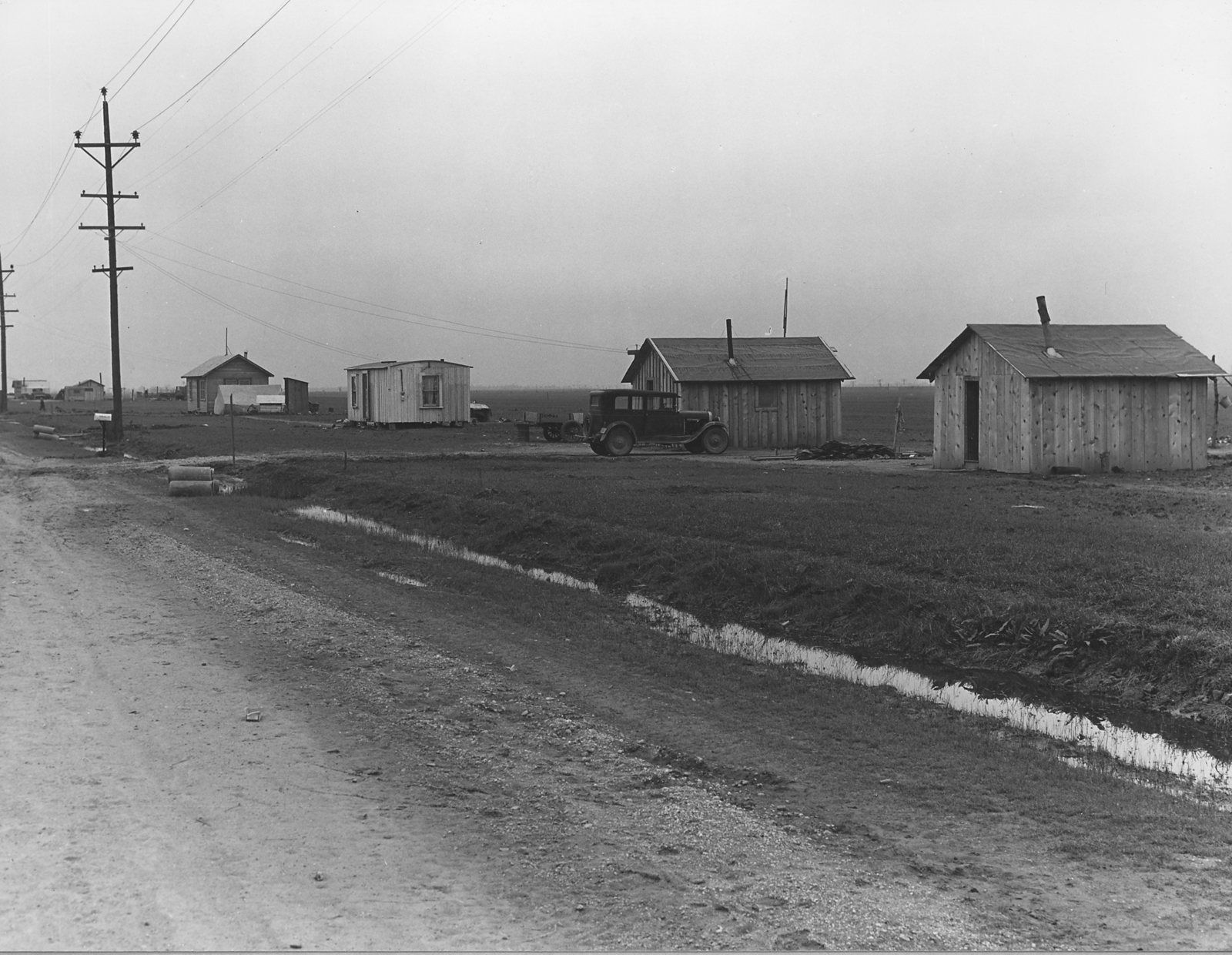 Black-and-white 1940 photo of newly built small wooden houses along a dirt road and drainage ditch on the flat farmland west of Olivehurst in Yuba County