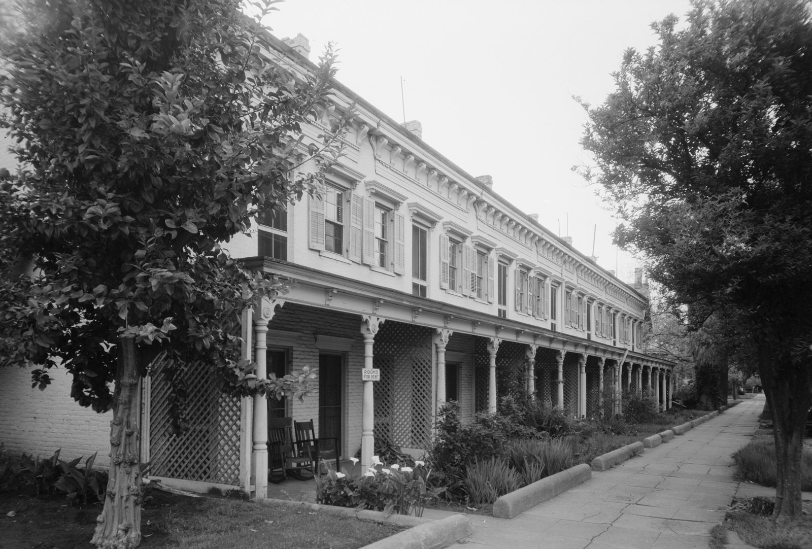 Black-and-white photo of a long two-story row of historic homes with a columned front porch and lattice, shaded by trees along C Street in Marysville, California