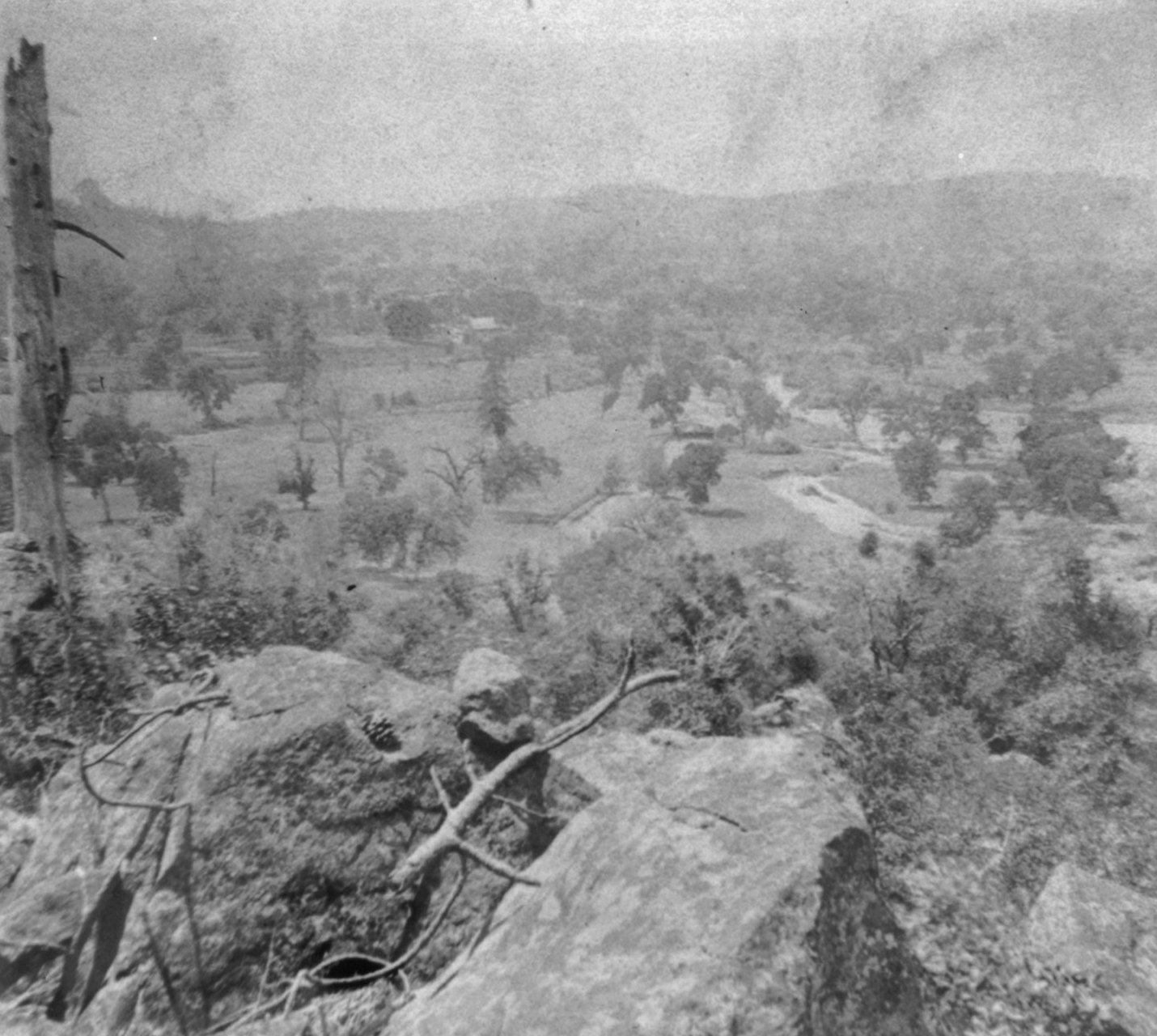 Black-and-white 1866 photo looking down from a rocky, brushy hillside over the open fields, oak trees and rolling hills of Penn Valley near Rough and Ready in Nevada County