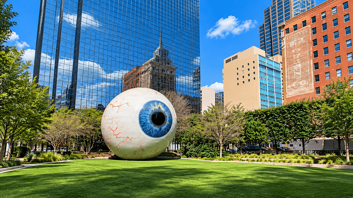 Giant blue eyeball sculpture by Tony Tasset in downtown Dallas across from The Joule, surrounded by lawn and city skyscrapers
