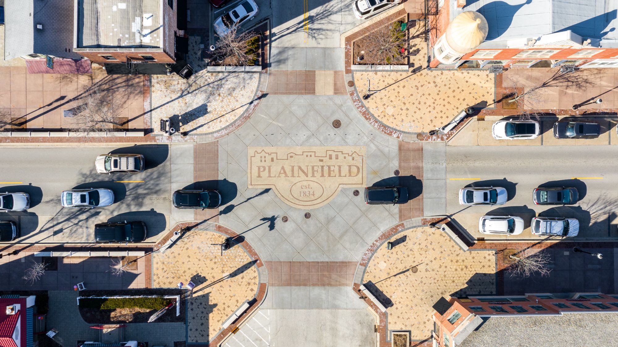 Overhead view of a downtown street intersection with a paved emblem reading Plainfield, est. 1834, framed by brick crosswalks, corner plazas and parked cars