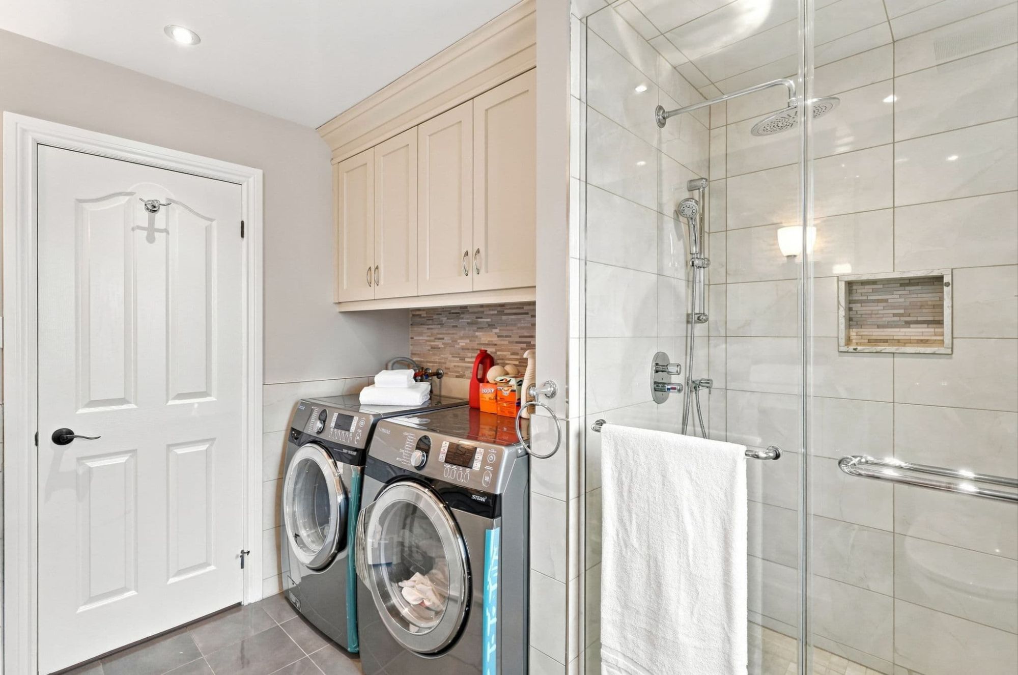 Residential laundry area with front-loading washer and dryer, built-in cabinets, folded towels and an adjacent glass shower