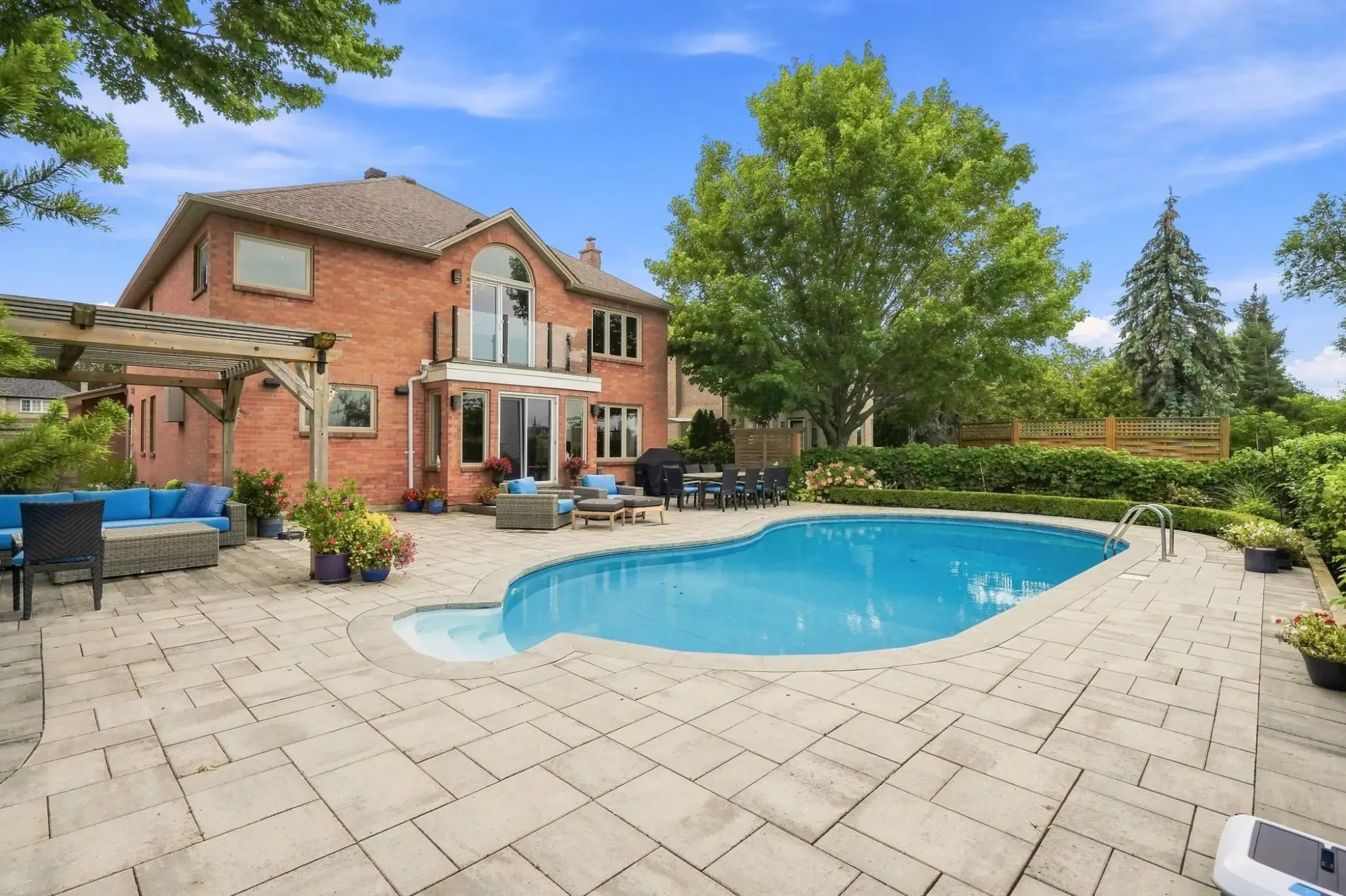 Two-storey brick home with a curved outdoor swimming pool, stone patio, pergola, outdoor seating and mature trees.
