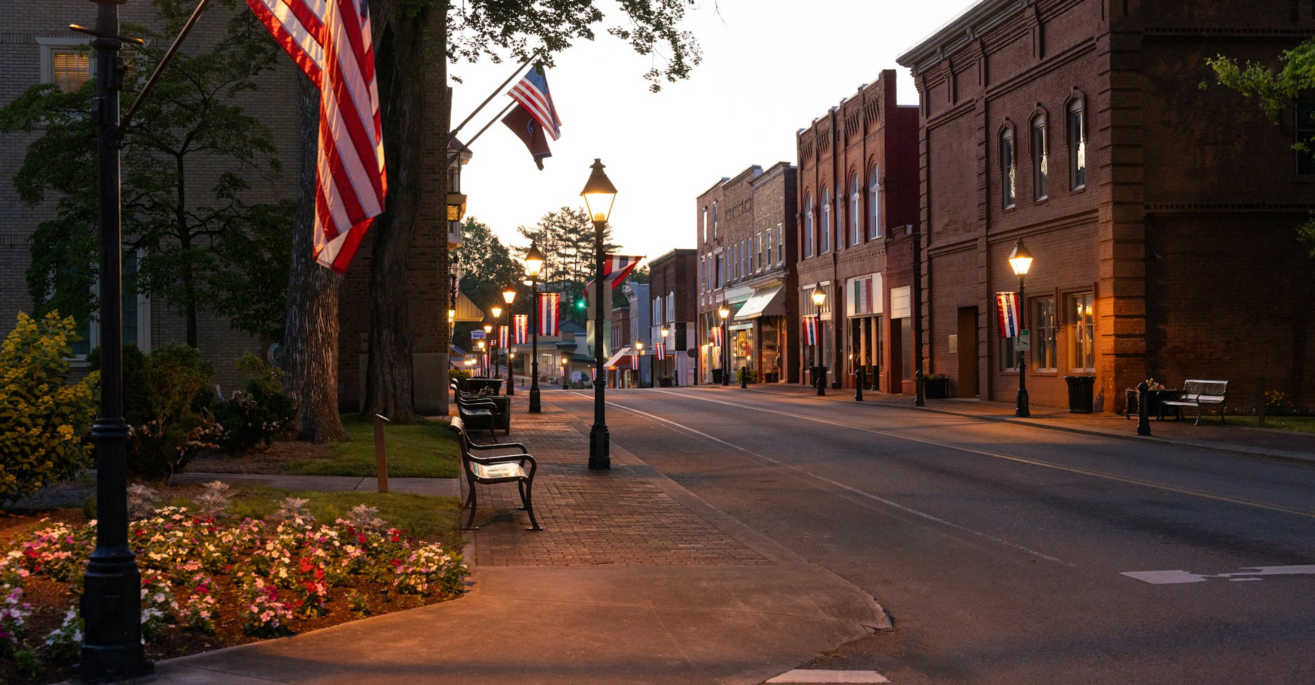 Brick storefronts, street lamps and flags along a small-town main street at dusk