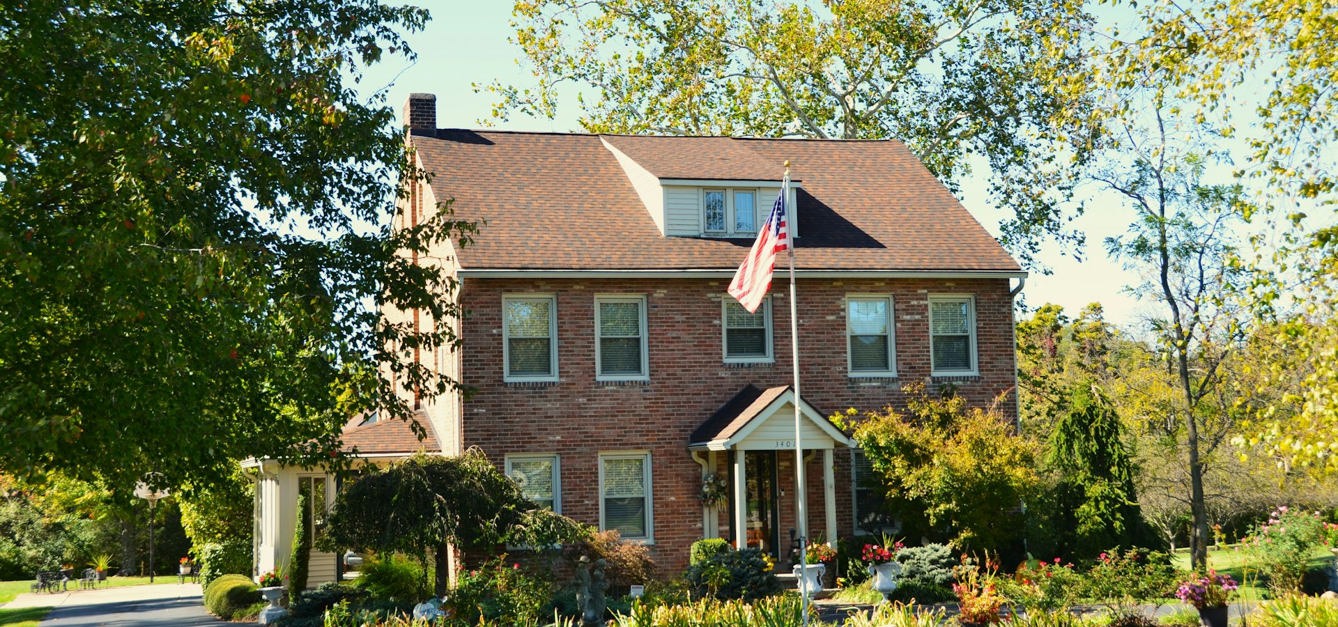 A brick two-story house with a covered entry and a garden, surrounded by trees in early autumn