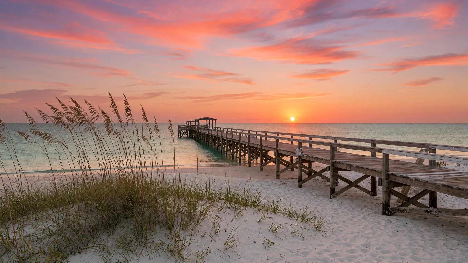 Golden hour sunset over the Alabama Gulf Coast with a pier extending into turquoise water, white sand beach, and sea oats in the foreground