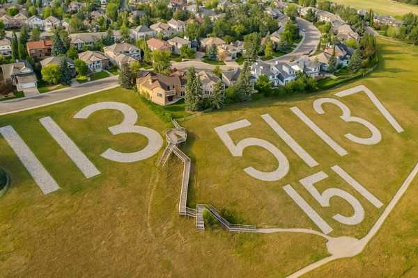 Aerial view of Battalion Park in Calgary, featuring large white stone numbers "113", "51", "137", and "151" embedded into a grassy hillside. A wooden staircase connects the hill to a suburban neighborhood with tree-lined streets and detached homes.