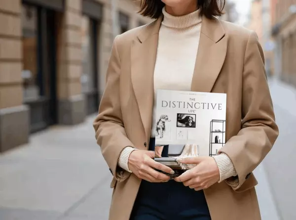 A person dressed in a stylish tan blazer and cream turtleneck sweater is holding a magazine titled "The Distinctive Life" in their arms. They are walking down a city street with blurred buildings in the background, conveying a sense of urban sophistication and modern lifestyle.