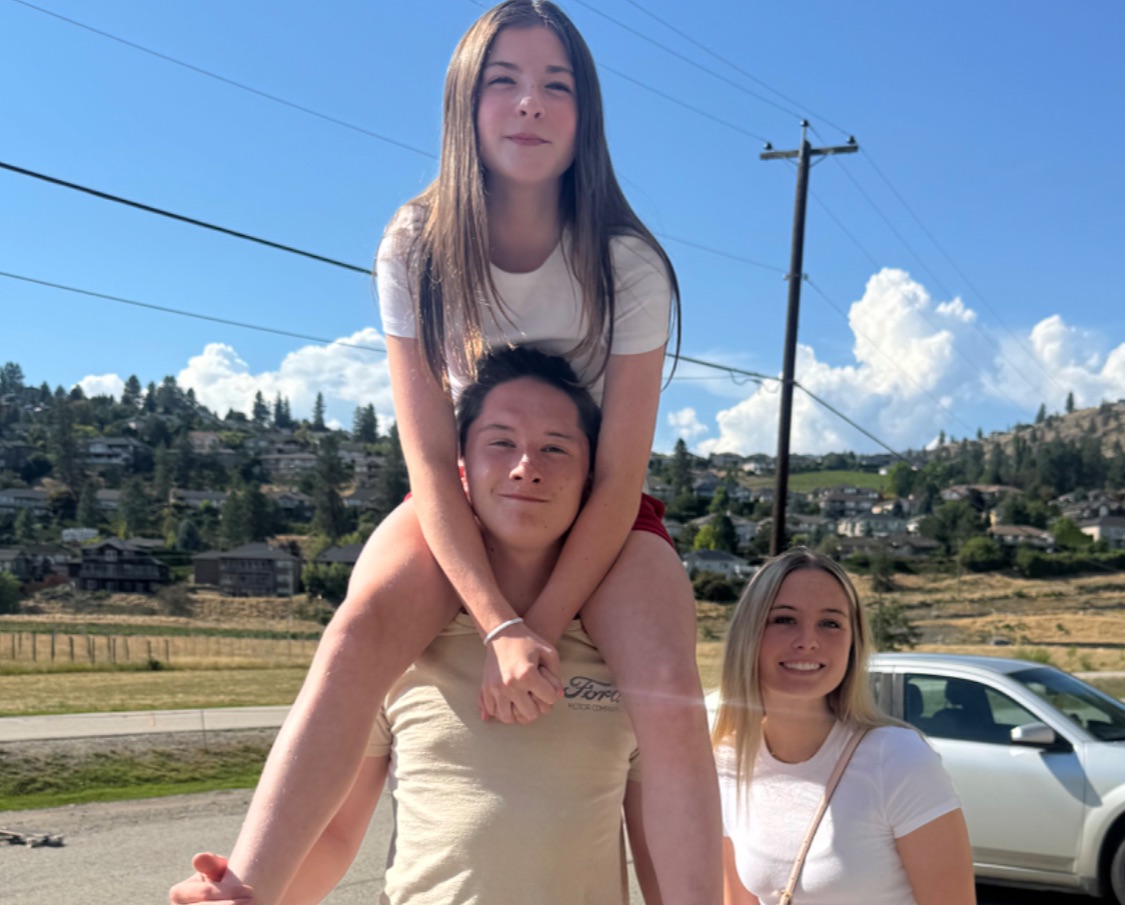 Outdoors on a sunny day, a teenage boy stands on the street carrying a younger girl on his shoulders while another teen stands beside them, all smiling. They’re dressed in light summer clothes with a backdrop of hills, houses, and a bright blue sky.