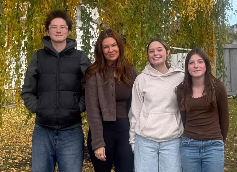 A woman, Laura Sikorski, stands outside with three children under a tree with golden fall leaves. They’re smiling and standing close together on a lawn covered in autumn leaves. The group is casually dressed in jackets, sweaters, and jeans.