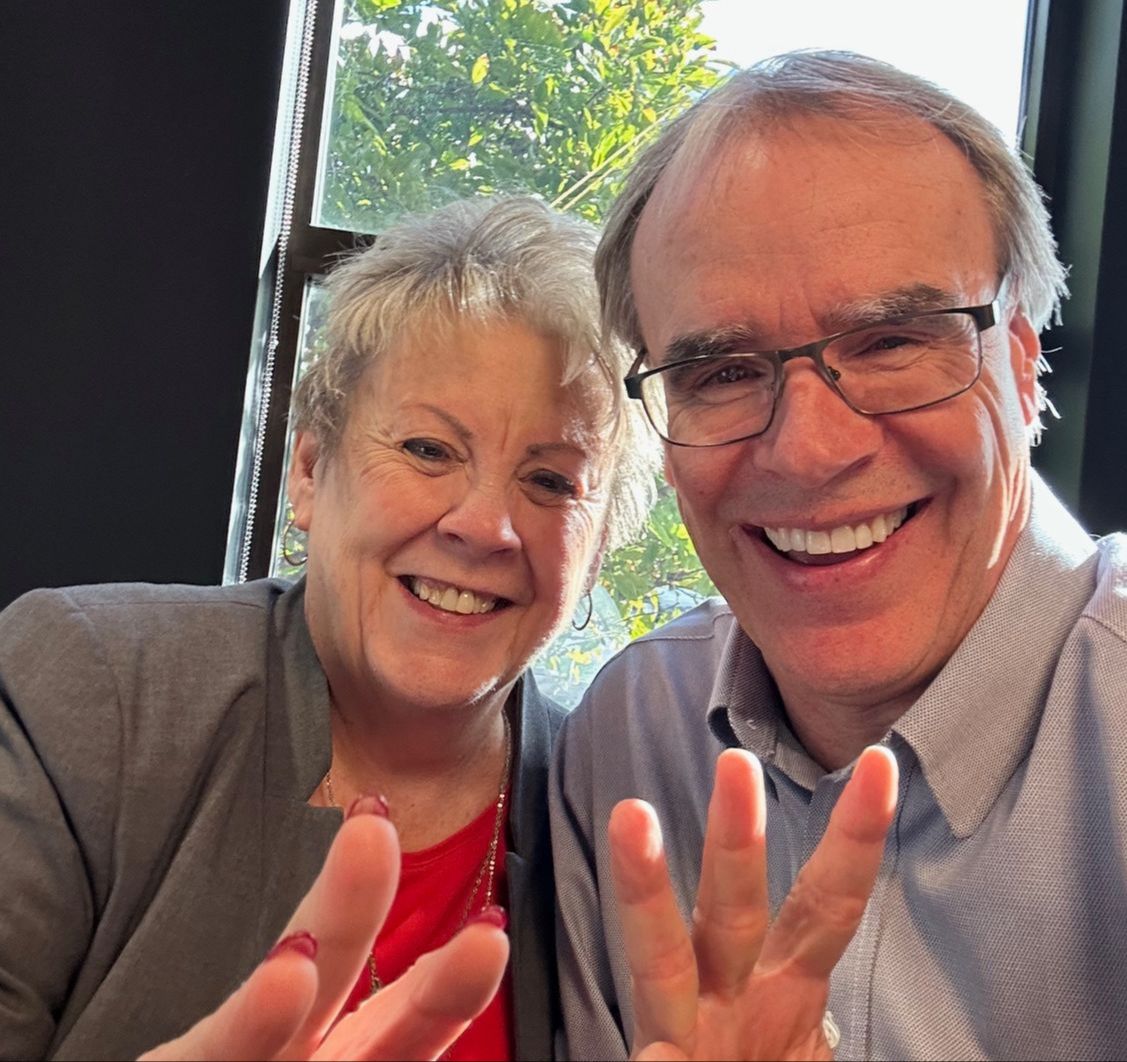 David Campbell, realtor, sits next to Phyllis as they hold up three fingers towards the camera, while smiling.