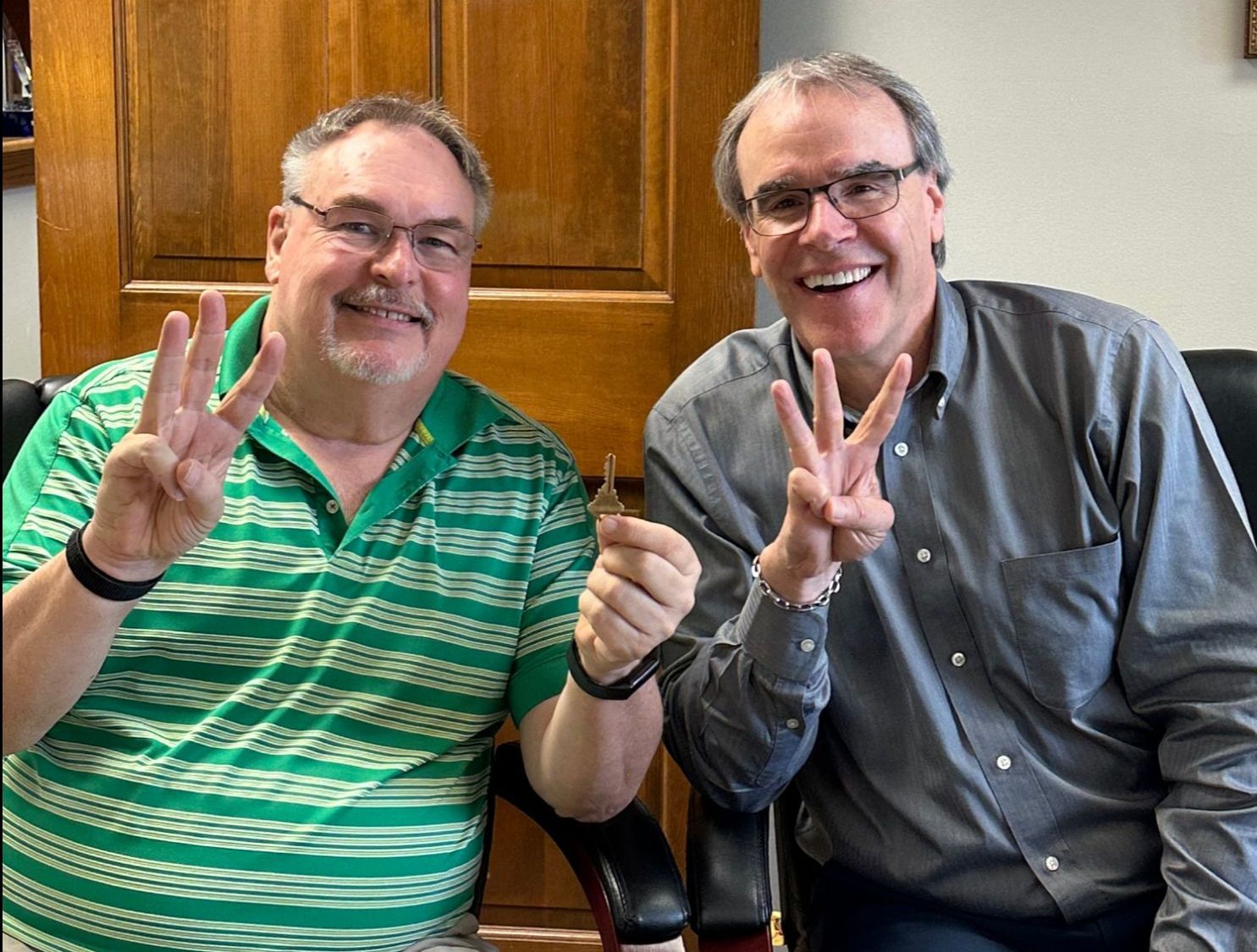 David Campbell, realtor, sits next to Mark as they both smile while facing the camera. Both of them are holding up three fingers and Mark has a house key in his left hand.