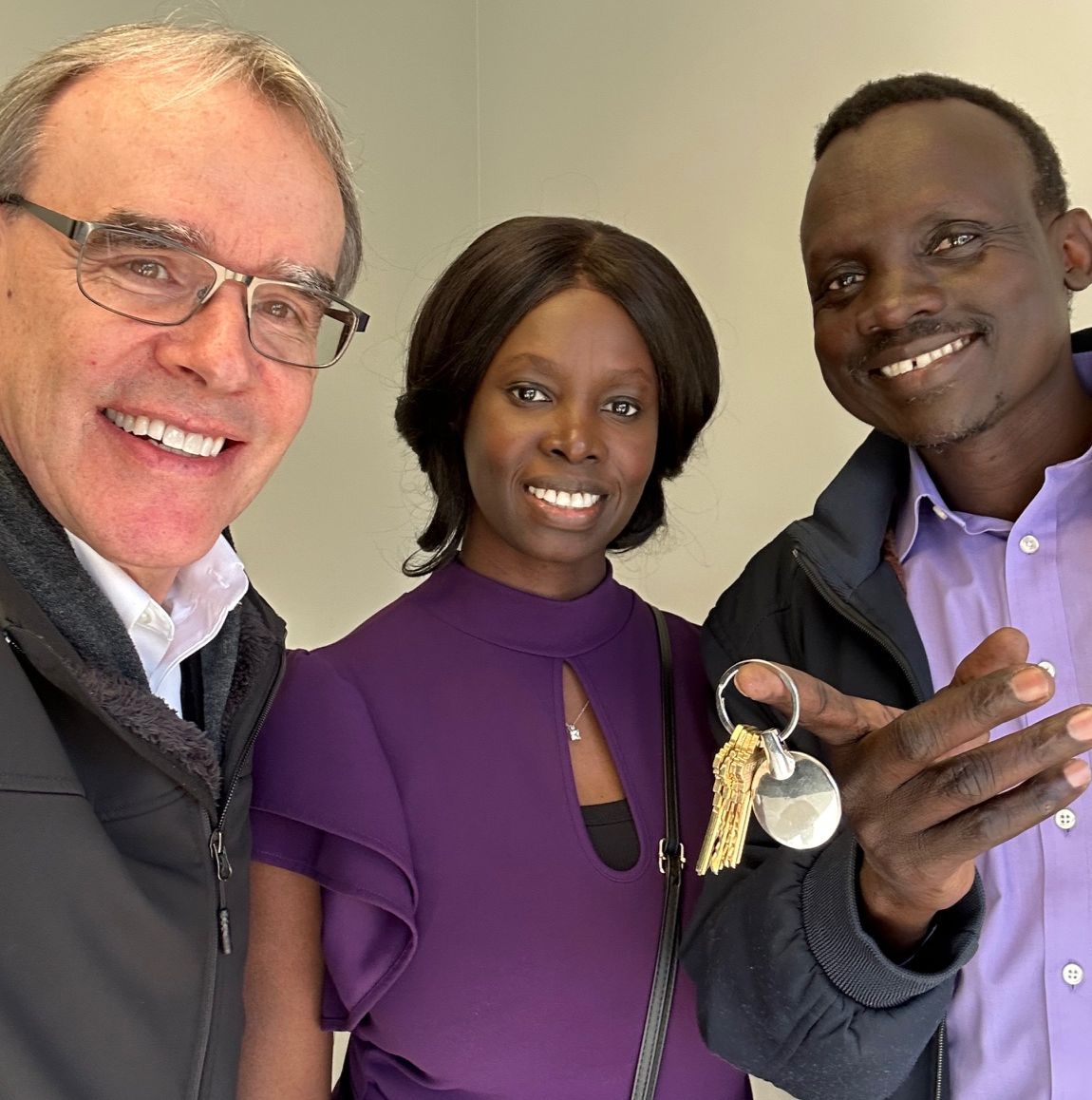 David Campbell, realtor, stands next to both Ngor and Abeny as they all face towards the camera, smiling. Abeny is holding a set of house keys.
