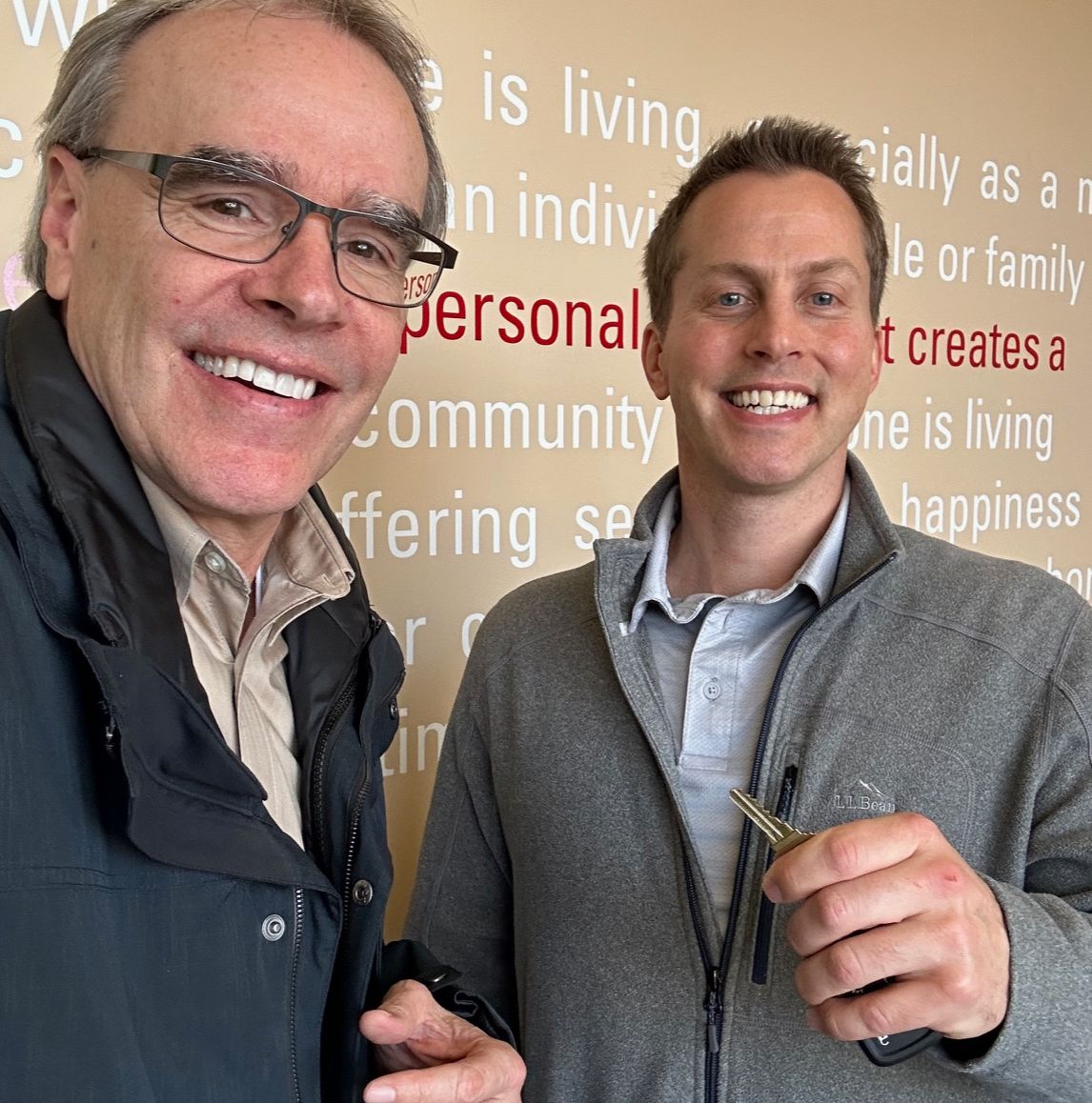 David Campbell, realtor, stands next to Erich, as they both face towards the camera, smiling. Erich is holding a house key.