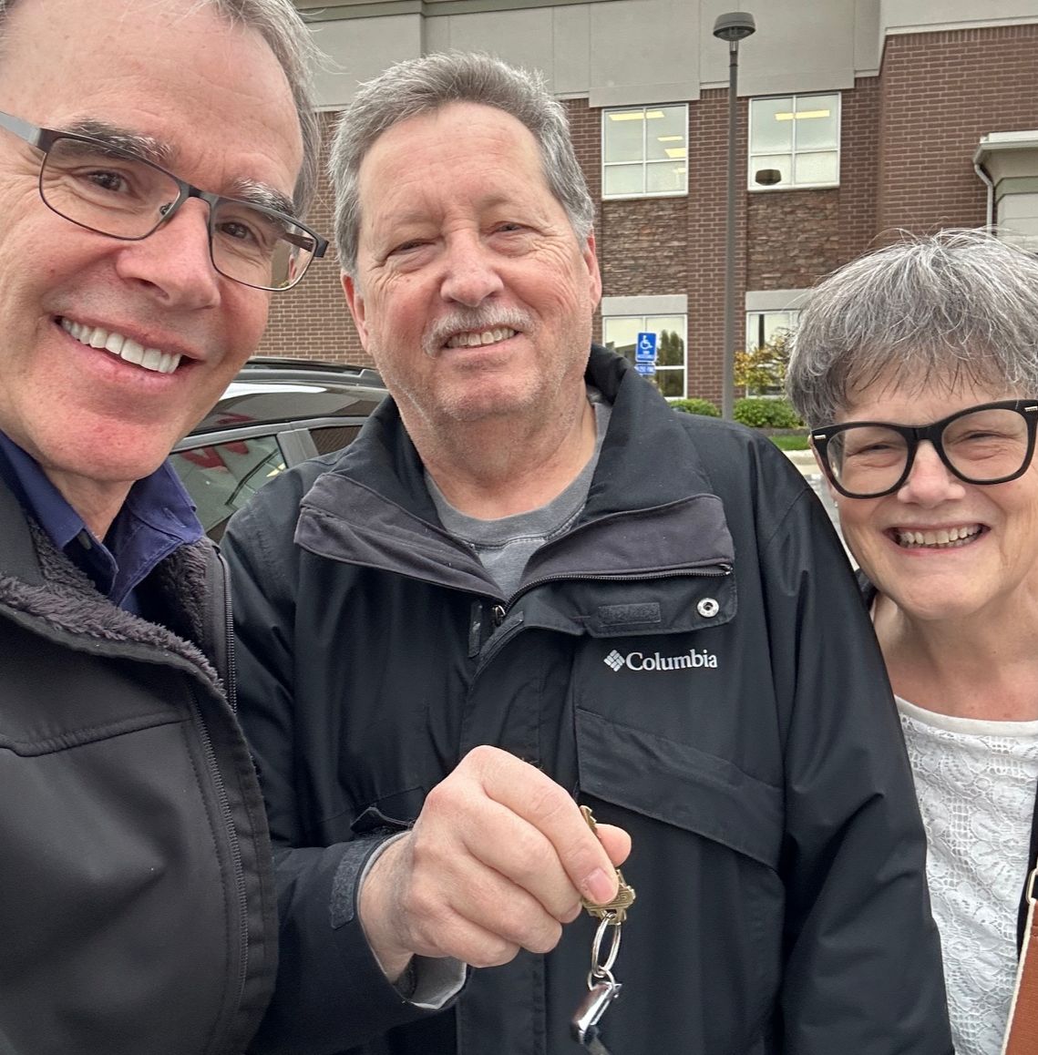 David Campbell, realtor stands next to both Doug and Nancy as they all face towards the camera, smiling. Doug is holding a house key.