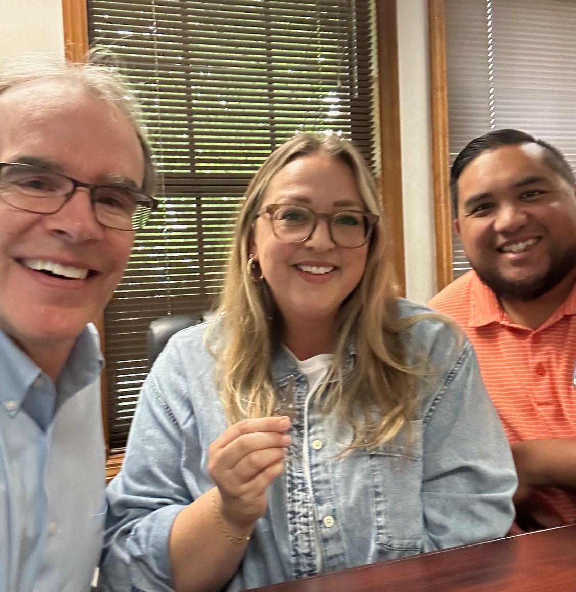 David Campbell sits next to both Katherine and Jorge as they all face towards the camera, smiling. Katherine is holding a house key.