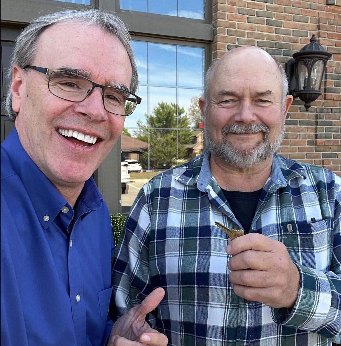 David Campbell realtor stands next to Robert outside. They're both facing towards the camera, smiling as Robert holds a house key.