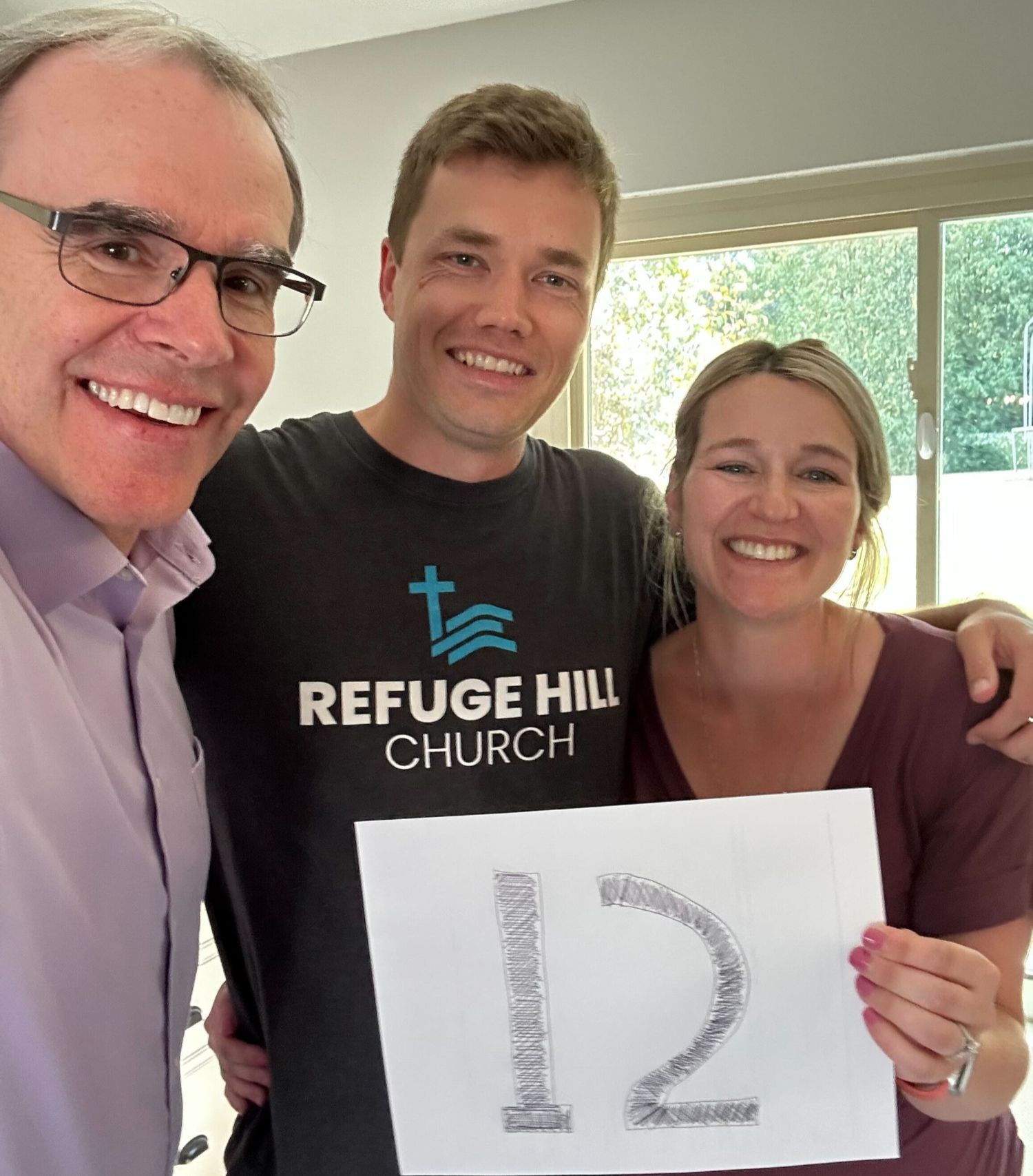 David Campbell, realtor stands next to both Nate and Liz as they all face towards the camera, smiling. Nate and Liz are holding up a sign that says "12".