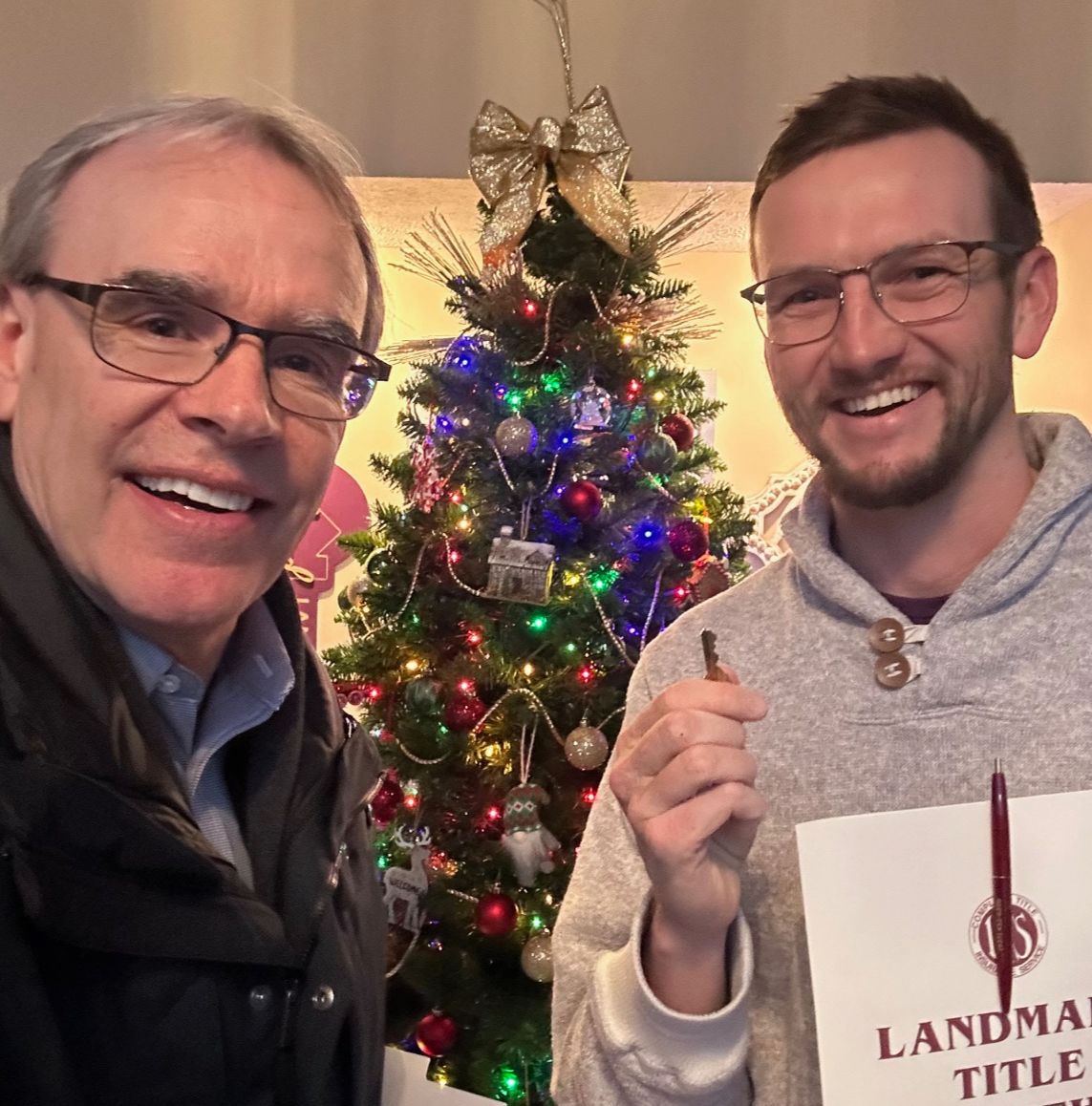 David Campbell, realtor stands next to Carl as they both smile towards the viewer. Clark is holding a house key and they're both standing in front of a lit Christmas tree.