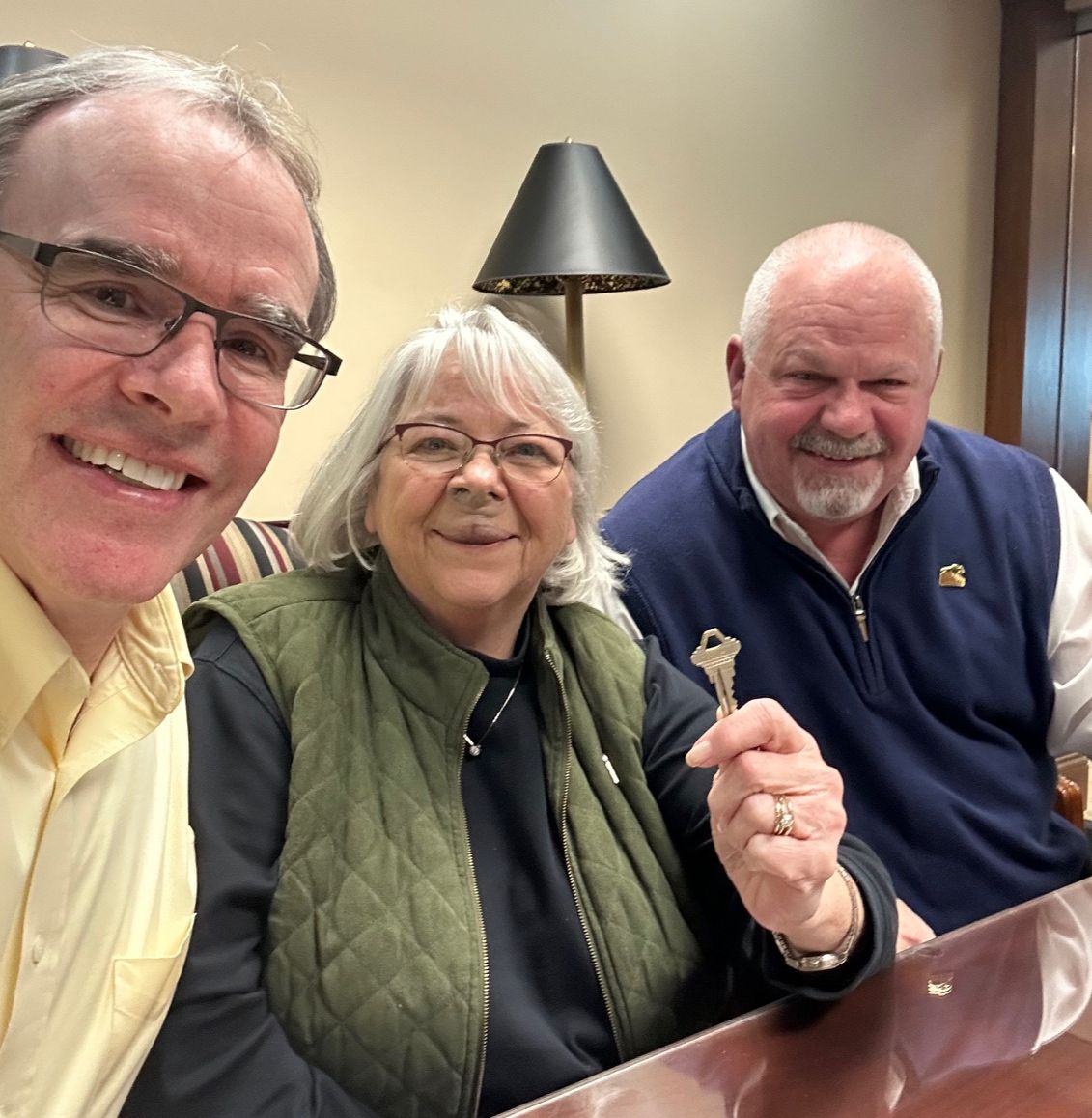 David is sitting to the left of a man and woman. The woman is holding his a house key - all of them are looking towards the camera smiling.