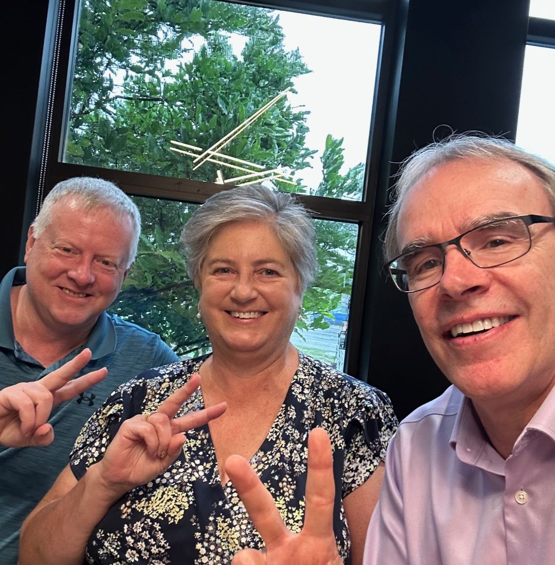 David sits next to a man and woman - all are facing towards the camera smiling, while holding up two fingers after a successful real estate transaction.