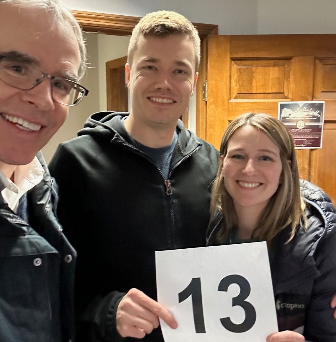 David Campbell, realtor stands next to both Nate and Liz as they all face towards the camera, smiling. Nate and Liz are holding up a sign that says "13".