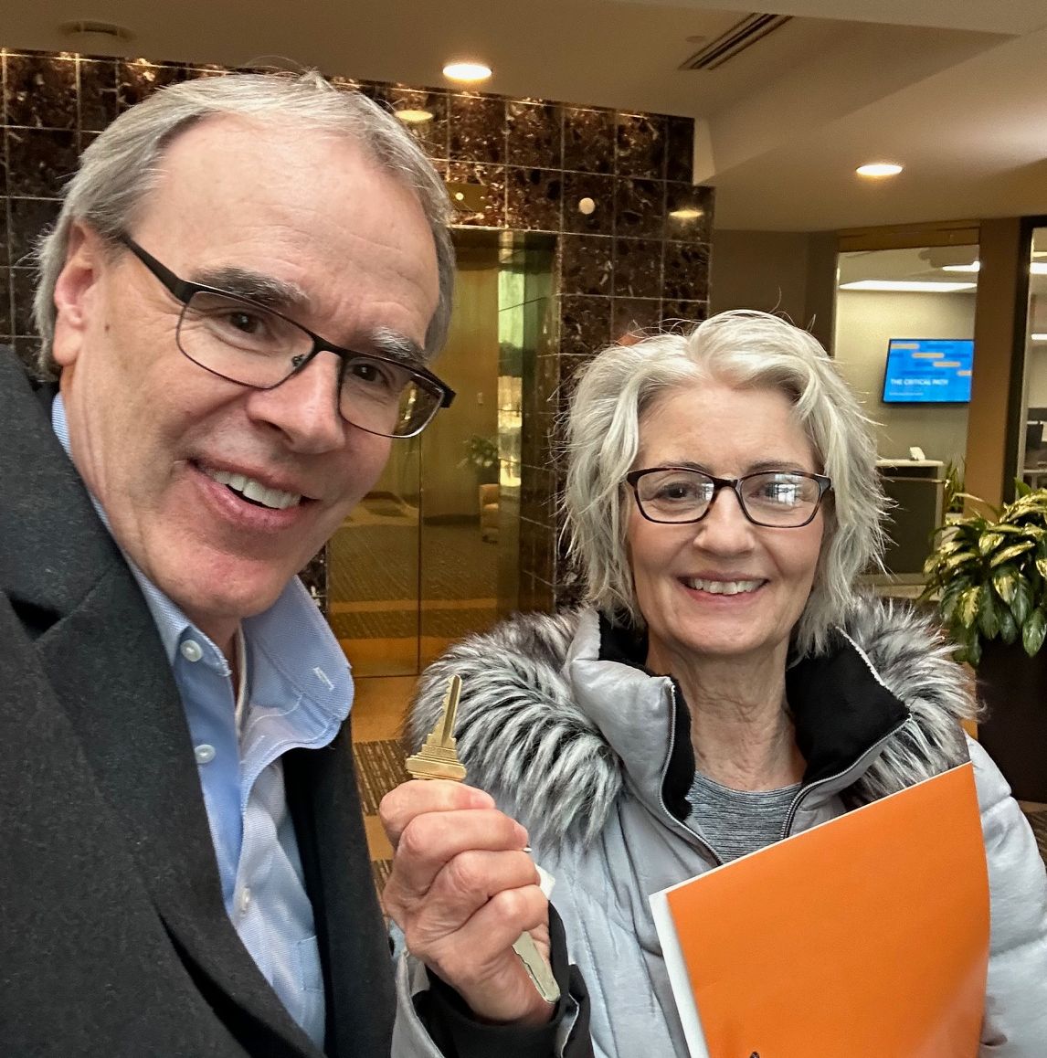 David Campbell, realtor stands next to Cindy as they both face towards the camera, smiling. Cindy is holding up a house key after a successful condo sale.