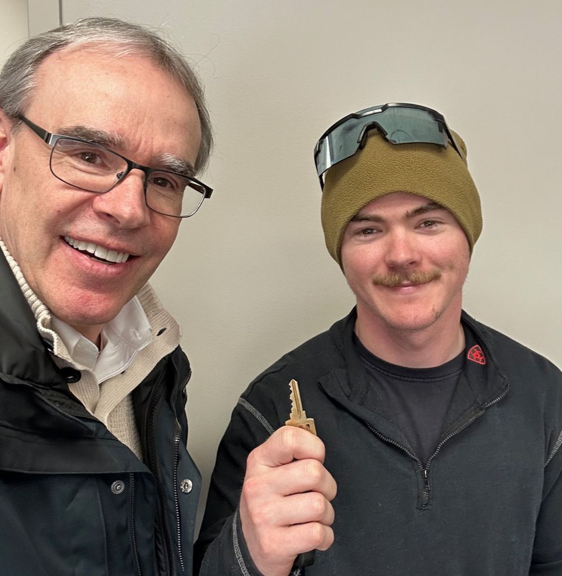 David Campbell, realtor stands next to Nick while they both face towards the camera, smiling. Nick is holding a house key after a successful home purchase.