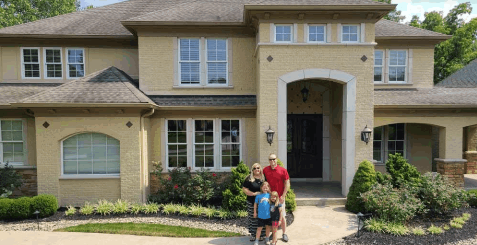 A happy family of four stands in front of a large house.