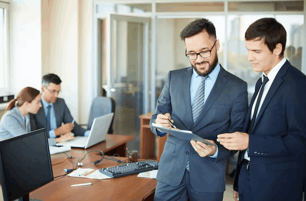 Stock Image of two men in suits looking over a clipboard. Two other office employees work in the background.
