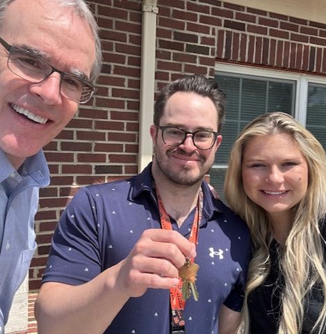 David Campbell, realtor, stands next to both Mike and Kate as they stand in front of their new home. Mike is holding house keys.