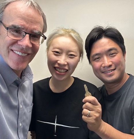 David Campbell, realtor, stands next to both Elliot and Jenny as they all face towards the camera, smiling. Elliot is holding a house key.