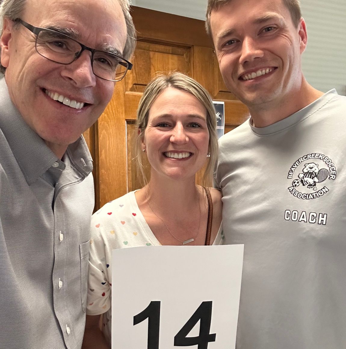 David Campbell, local realtor, stands next to both Nate & Liz as they hold up a paper that has the number 14 on it.