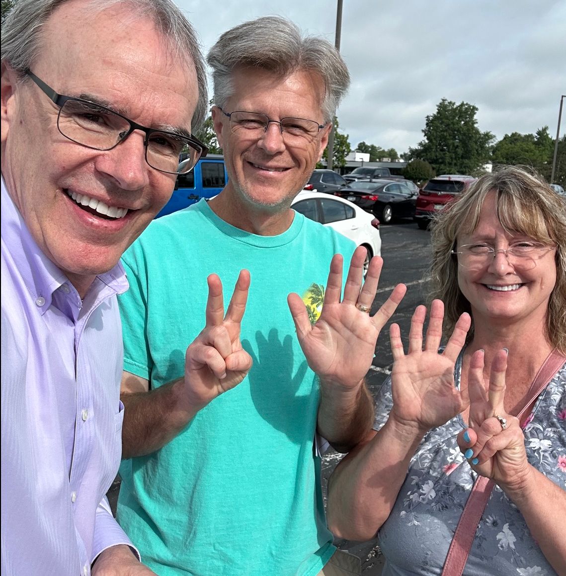 David Campbell, realtor, stands next to both David and Sandy in a parking lot. David and Sandy are holding up seven fingers each.